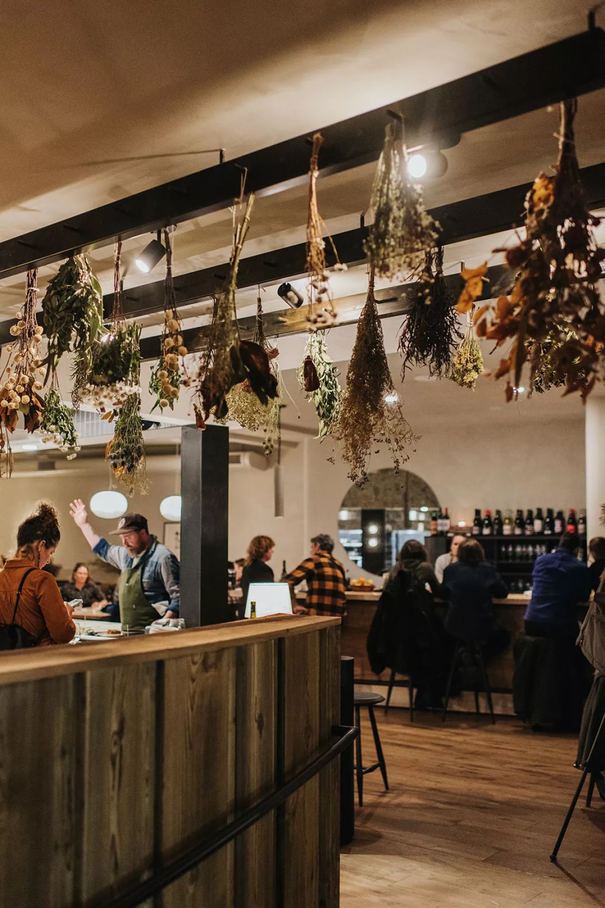 Chleo at night — dried herbs hanging from a steel beam, room full of guests at the bar and tables, chef gesturing