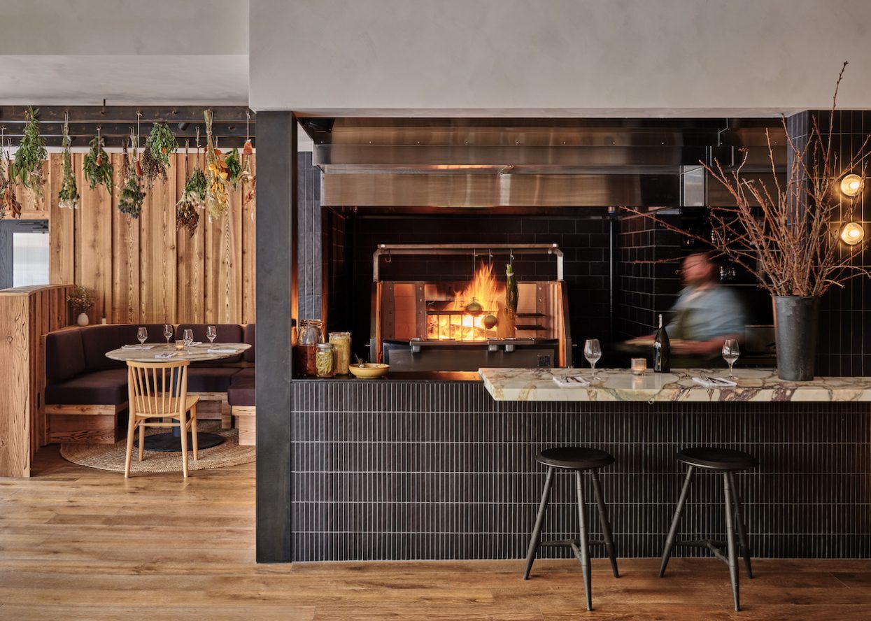 The wood-fired hearth at Chleo — open kitchen, dried herbs hanging on spalted maple, terrazzo bar