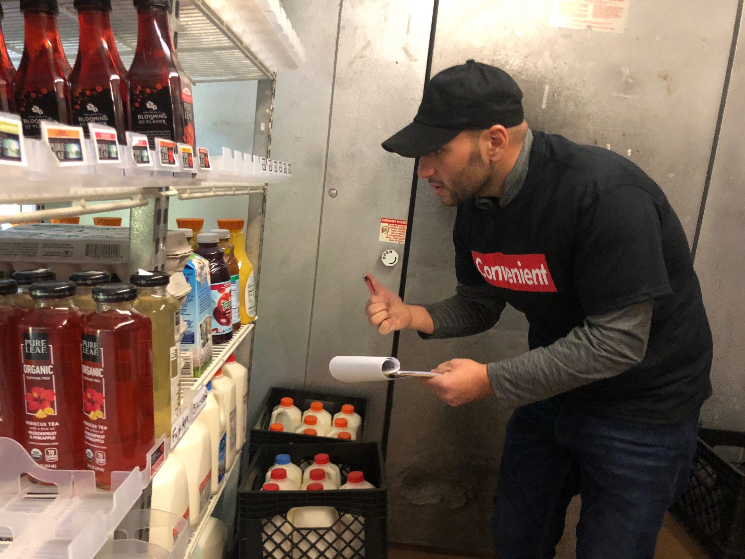 Khalil Jamal, Convenient Deli's current owner and a SUNY New Paltz graduate, in a black cap and Convenient Deli shirt at the walk-in cold case, clipboard in hand