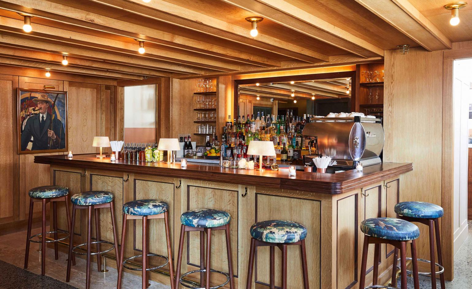 The bar at Hotel Kinsley — oak panelling, brass-trimmed bar, blue floral barstools, framed art on a wood-panelled wall