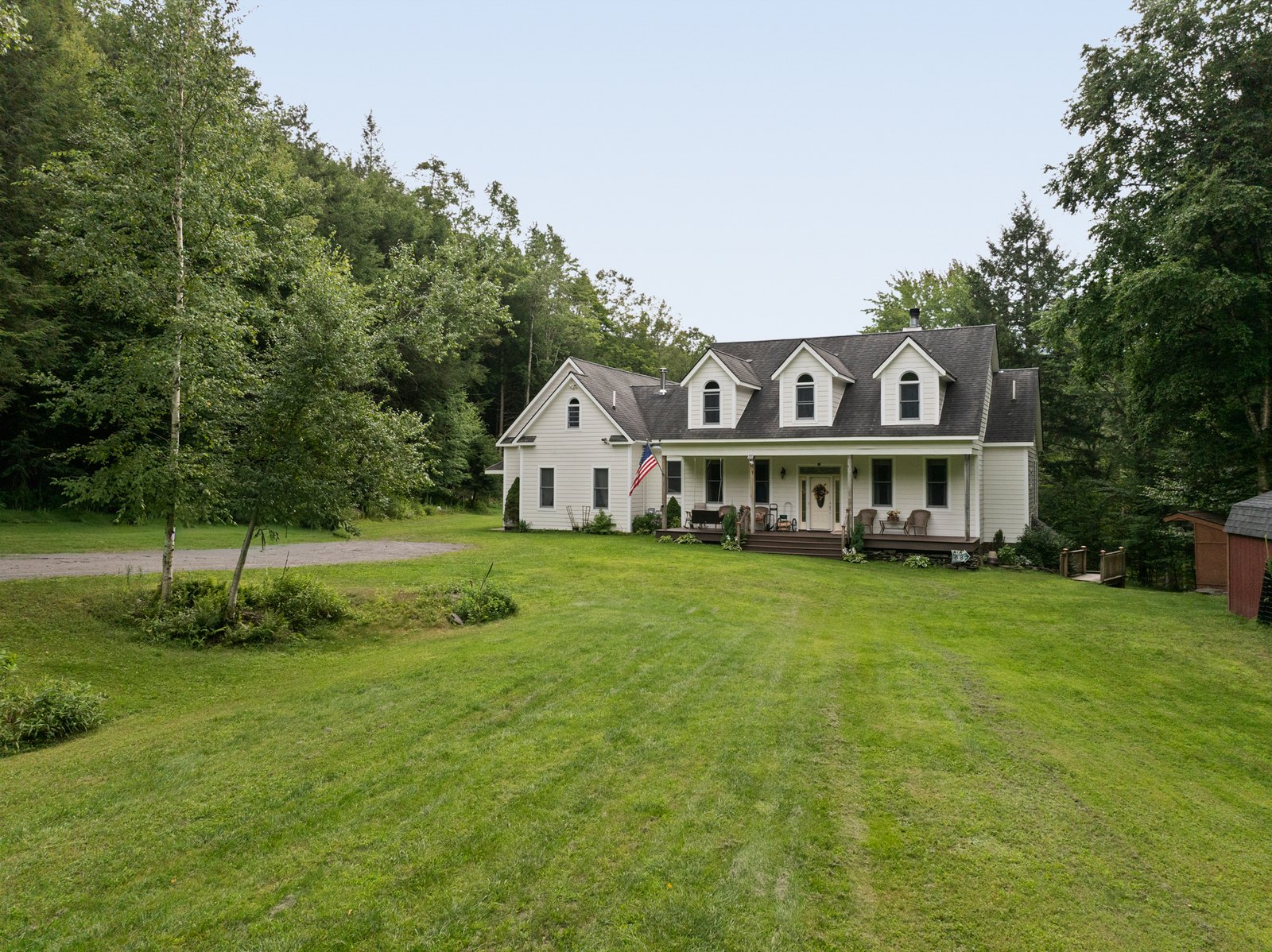 A white gabled farmhouse-style house across an open lawn edged by forest.
