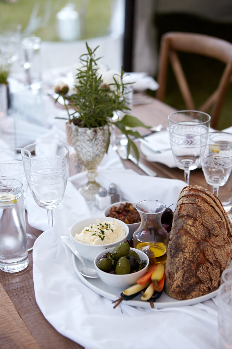 A wedding-tent charcuterie board catered by Main Course — housemade bread, whipped ricotta, olives, olive oil, cudighi at a linen-set farm table with rosemary centerpiece