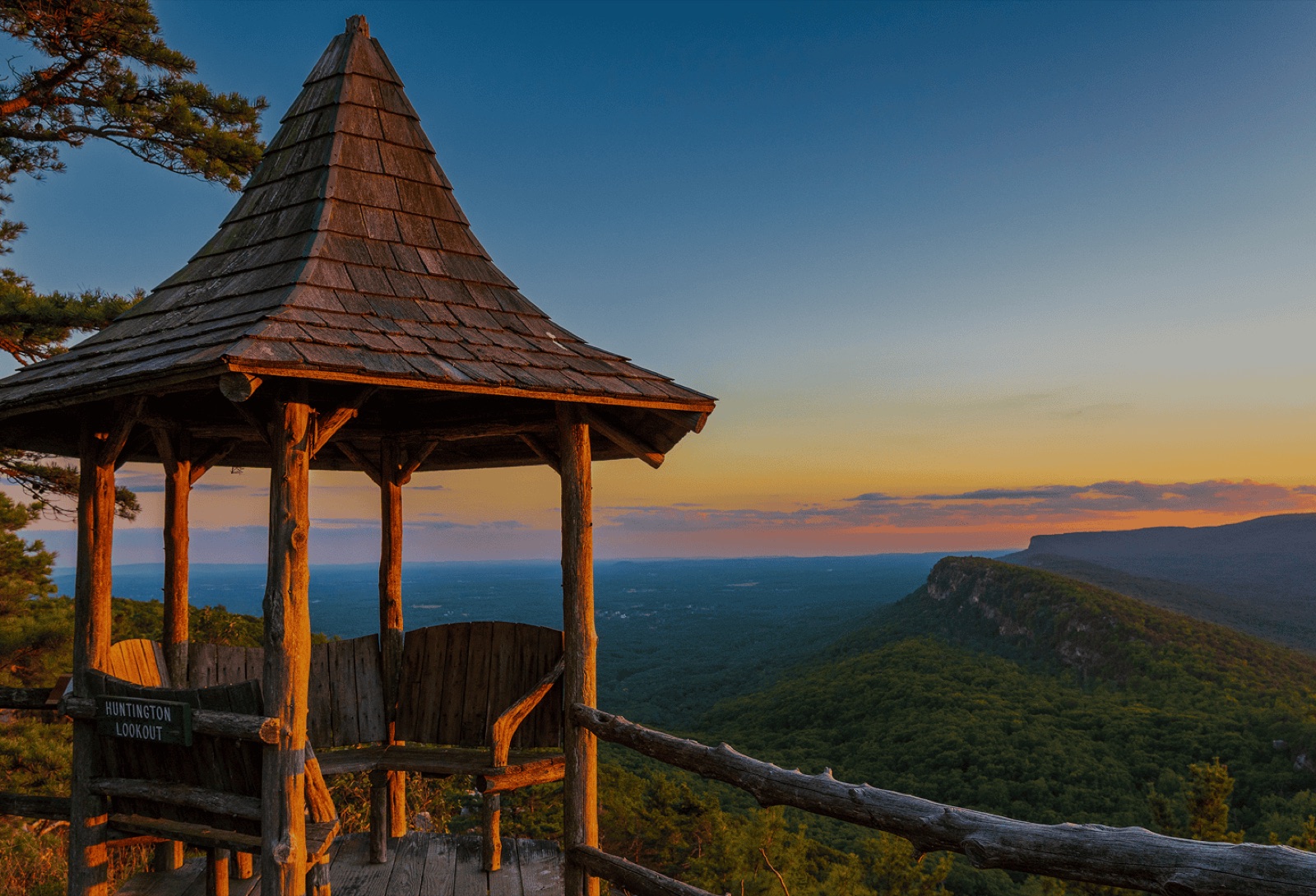 A Mohonk hiking-trail gazebo at sunset — wood structure with bench, looking out over the Shawangunk Ridge with golden-hour light on the cliffs and Catskill foothills beyond