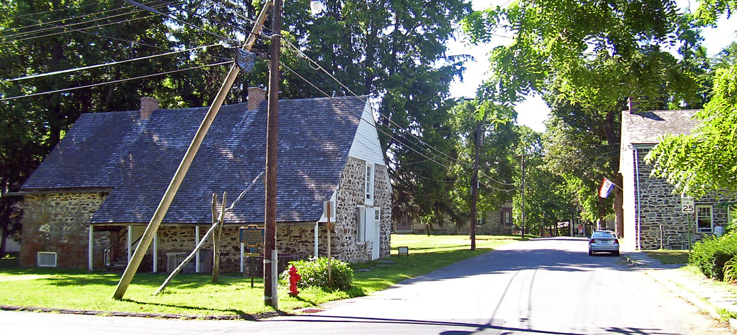Historic Huguenot Street in New Paltz — early-1700s stone houses set back from the street under summer trees, weathered slate roofs, the Wallkill River neighborhood beyond