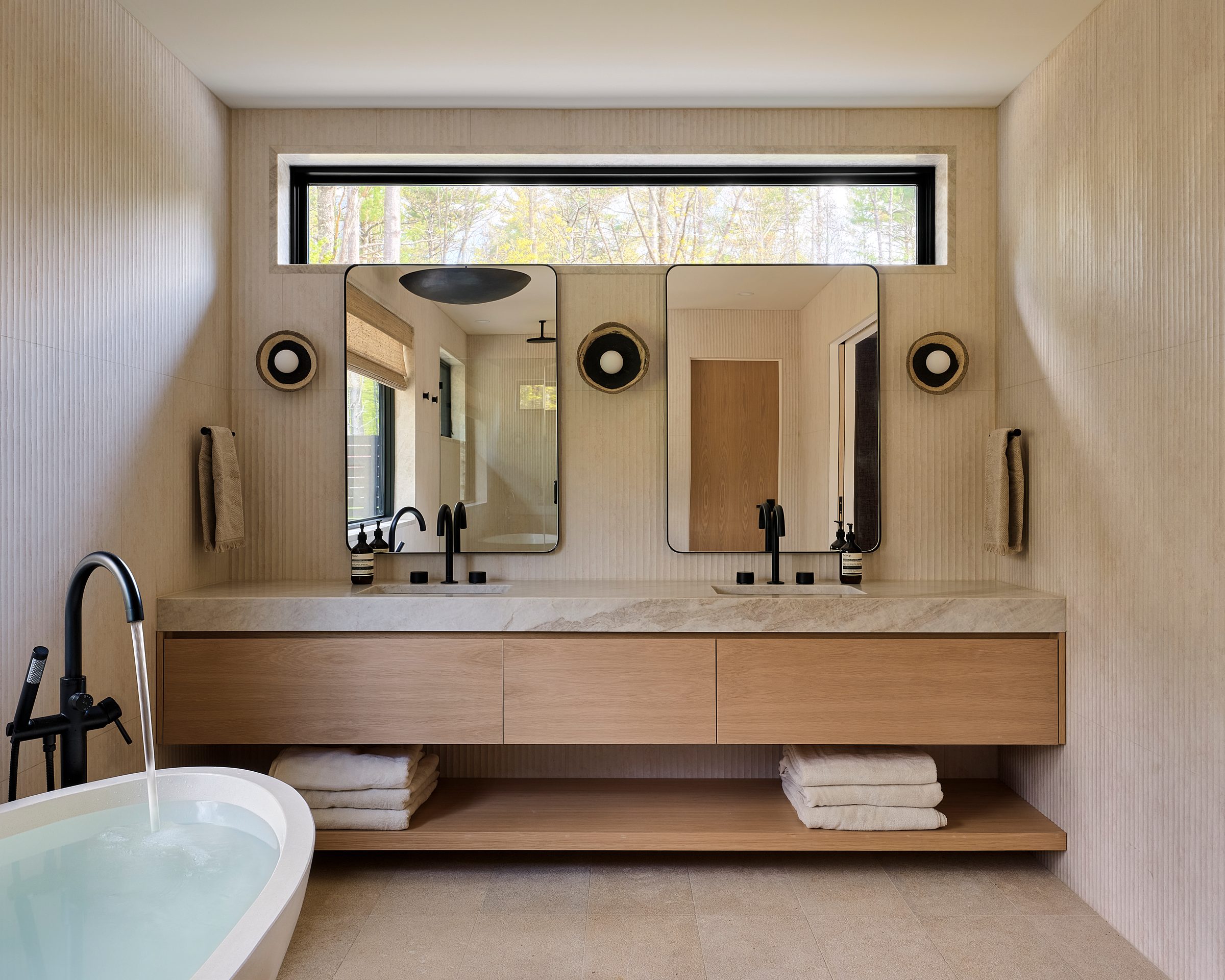 A bath with a freestanding tub, a floating pale-oak double vanity and a clerestory window.