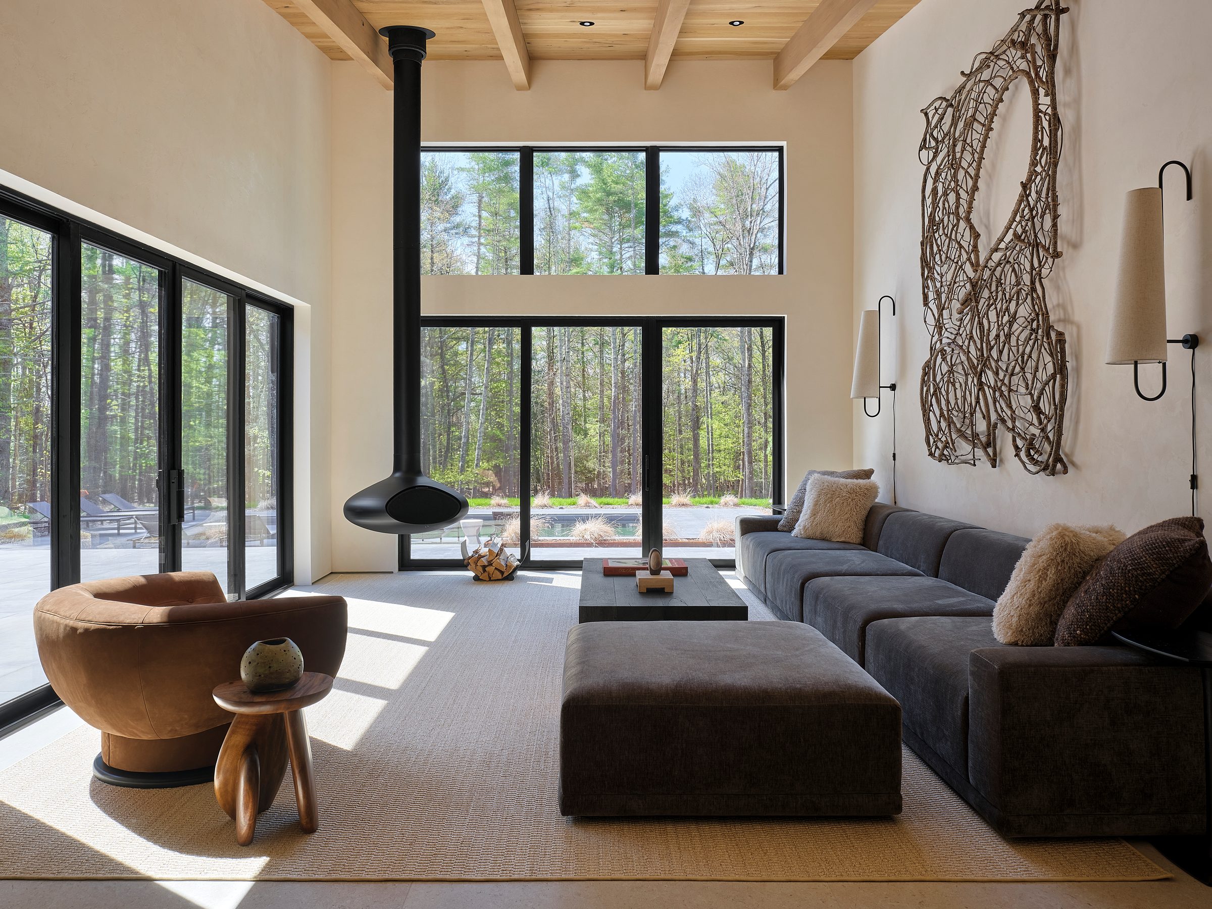 A double-height living room with a suspended round fireplace and tall black-framed windows to the woods.