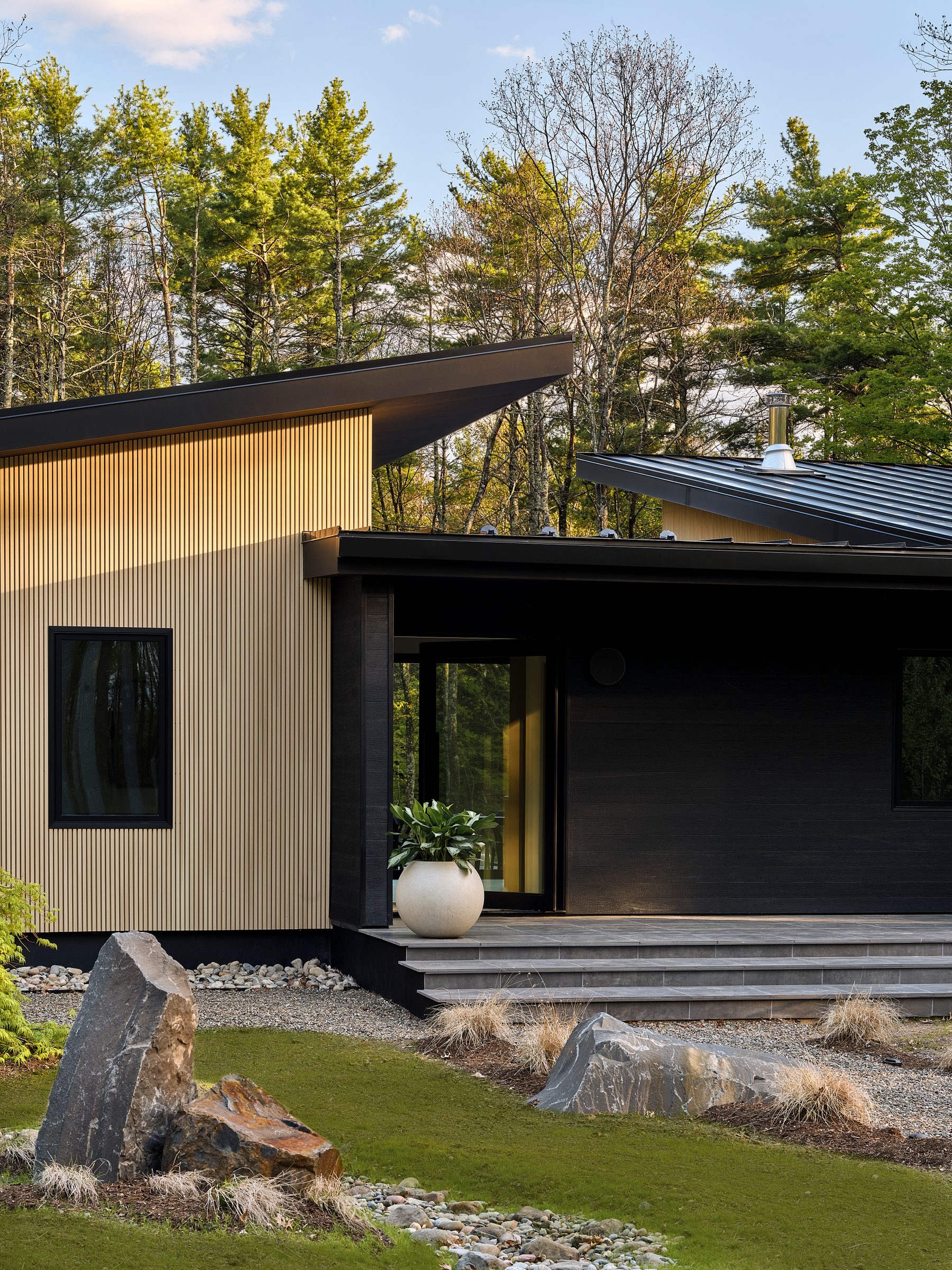 The front of the house: pale cladding, black trim and a shallow dark roof among pines.