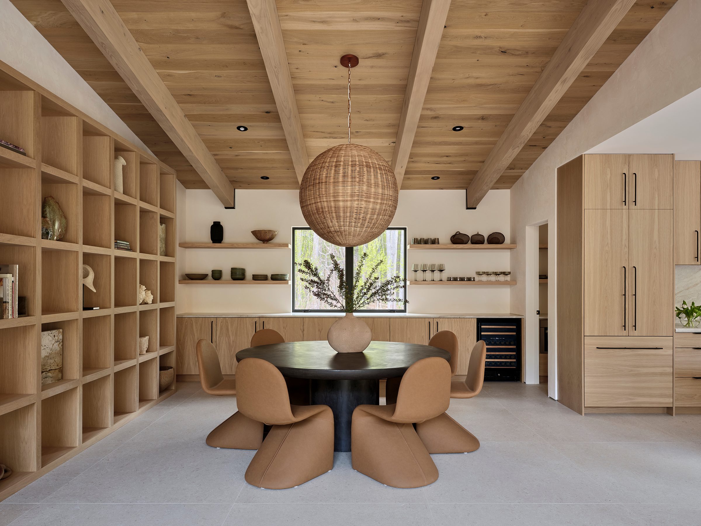 A dining area with a round table, built-in shelving and a woven pendant under a beamed ceiling.