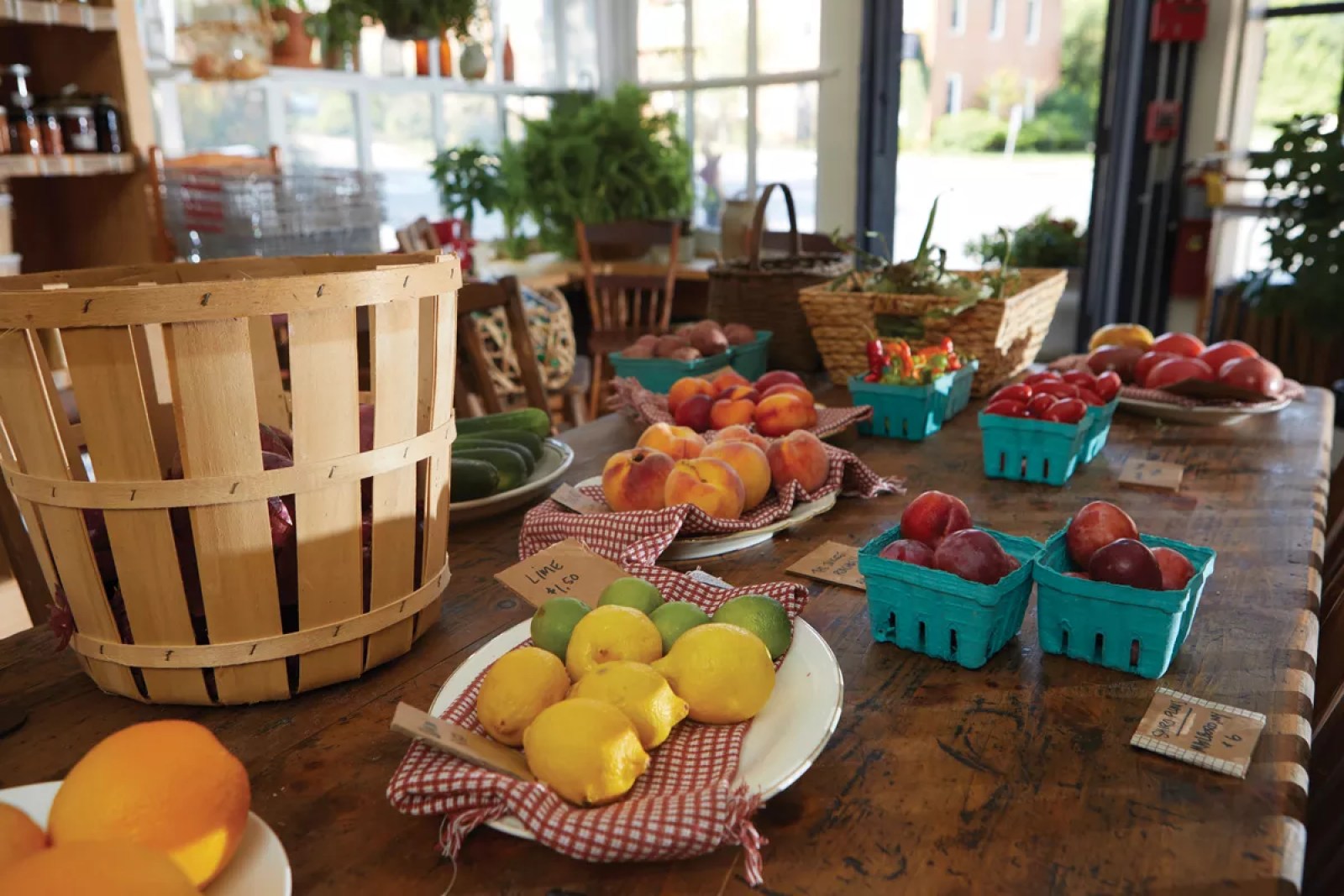 A farm table at the front of Rosie General — peaches, plums, lemons and limes on vintage china, blue cardboard berry pints, woven baskets — sunlight pouring in through the mullioned corner windows